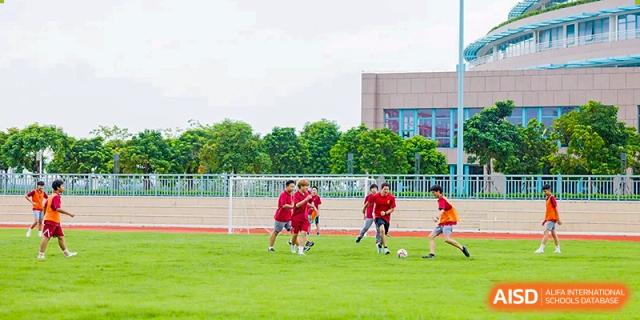 A group of students in red jerseys playing a soccer match on a large green field with a goalpost in the background at Guangzhou ULink International School.