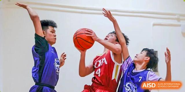 A student athlete in a red jersey focuses intently while going for a layup during a basketball game in the gymnasium at Guangzhou ULink International School.