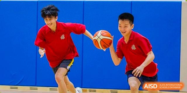 Two students in red shirts practice dribbling and passing a basketball against a blue padded wall in the school gym at Guangzhou ULink International School.