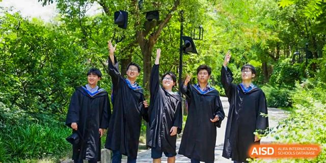 Three graduates in black gowns cheer and toss their caps into the air under green trees at Yew Wah International Education School of Guangzhou