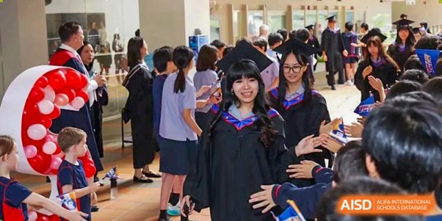 A smiling female graduate in a black gown walks through a crowd of fellow students during a graduation event at Yew Wah International Education School of Guangzhou