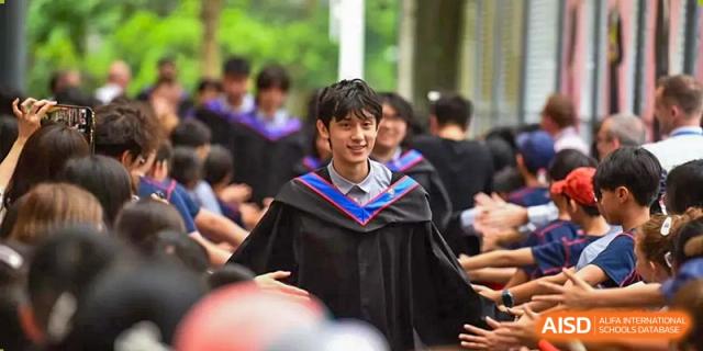A young male graduate in a black academic gown smiles while standing among his peers during a school ceremony at Yew Wah International Education School of Guangzhou