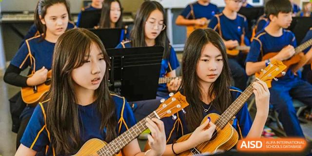 Three female students in blue uniforms sit together and play ukuleles during a school music performance or class at Yew Wah International Education School of Guangzhou