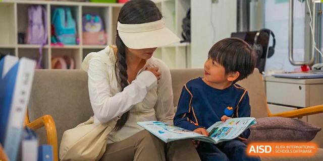A woman in a white sun visor sits with a young boy and looks at a book together during an outdoor activity at Yew Wah International Education School of Guangzhou