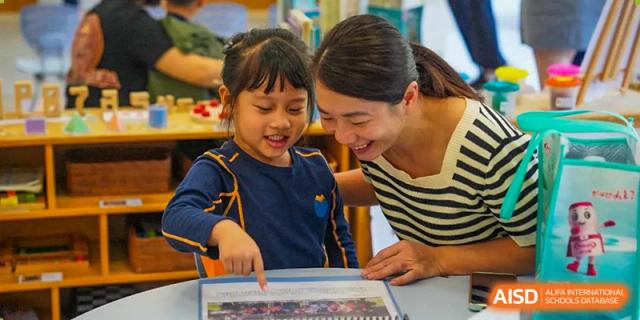 A smiling teacher leans over a desk to help a young girl with her schoolwork in a friendly classroom setting at Yew Wah International Education School of Guangzhou