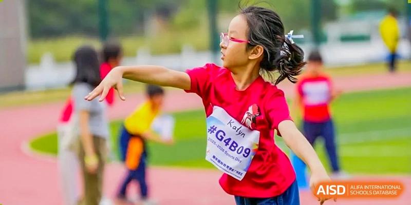 A female student in a red shirt with an identification bib prepares to throw a shot put on an outdoor field at Yew Wah International Education School of Guangzhou