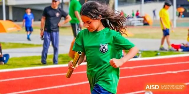 A young girl in a green shirt runs on a track while holding a golden relay baton during a sports competition at Yew Wah International Education School of Guangzhou
