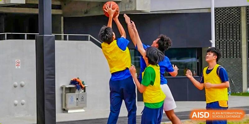 Three students in colorful sports vests compete for a basketball under the hoop during an outdoor game at Yew Wah International Education School of Guangzhou