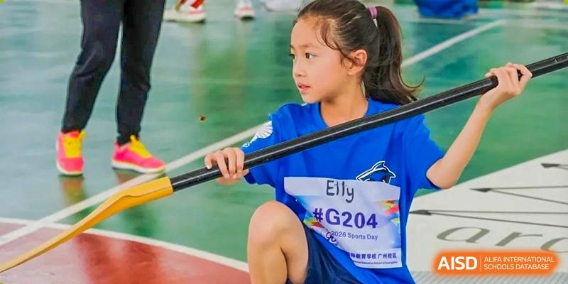 A young girl in a blue shirt holds a long black athletic pole while kneeling on an outdoor track surface at Yew Wah International Education School of Guangzhou