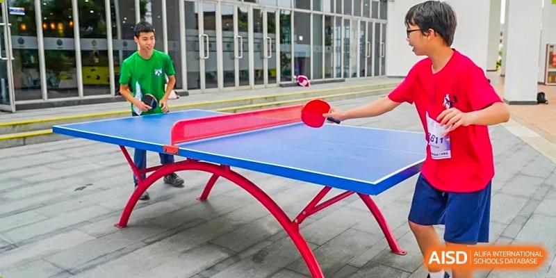 A hand holds a red table tennis paddle ready to hit a ball across a blue outdoor ping pong table at Yew Wah International Education School of Guangzhou