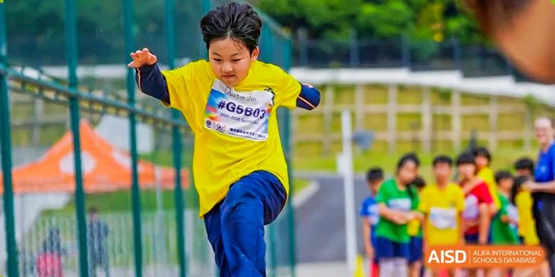 A young boy in a yellow sports shirt and blue shorts jumps over a hurdle on an outdoor track at Yew Wah International Education School of Guangzhou