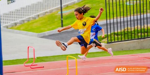 A young girl in a yellow shirt leaps over a yellow hurdle during a track and field event at Yew Wah International Education School of Guangzhou