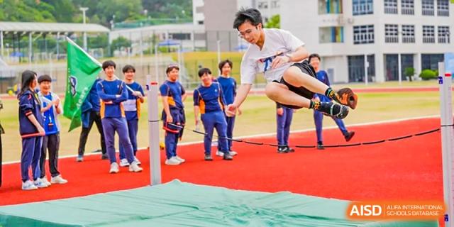 A male student in a white shirt performs a high jump over a bar onto a green landing mat at Yew Wah International Education School of Guangzhou