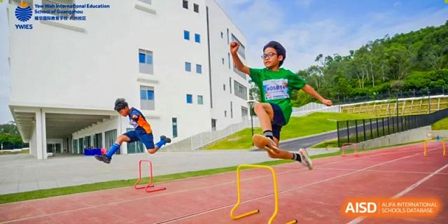 A student in a green shirt and glasses jumps over a yellow hurdle on an outdoor track at Yew Wah International Education School of Guangzhou