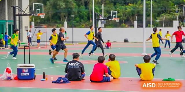 Students in sports uniforms play on an outdoor basketball court with a green forested background at Yew Wah International Education School of Guangzhou