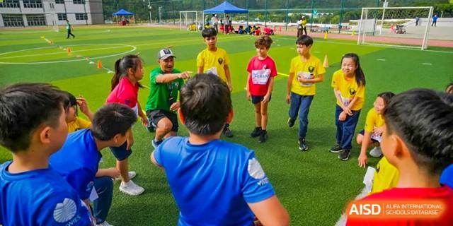 A group of students in various colored sports shirts listen to a coach on a green athletic field at Yew Wah International Education School of Guangzhou