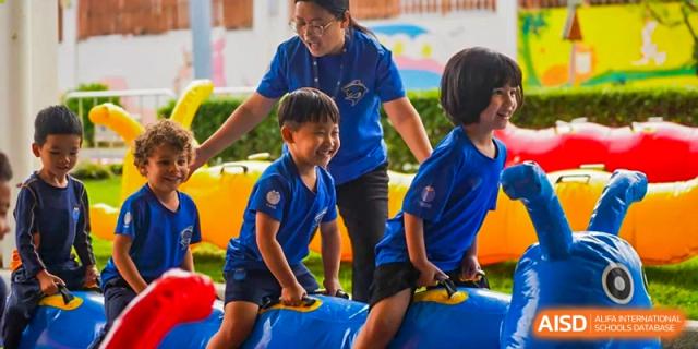 A teacher helps a young student in a blue shirt balance on an inflatable blue play structure outdoors at Yew Wah International Education School of Guangzhou