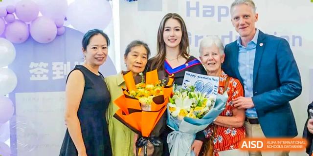 A female graduate in a black and blue gown holds large bouquets of yellow flowers with her family at Yew Wah International Education School of Guangzhou
