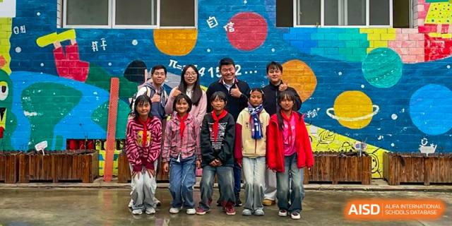 A group of students and teachers pose for a photo in front of a colorful mural on an outdoor wall at Yew Wah International Education School of Guangzhou