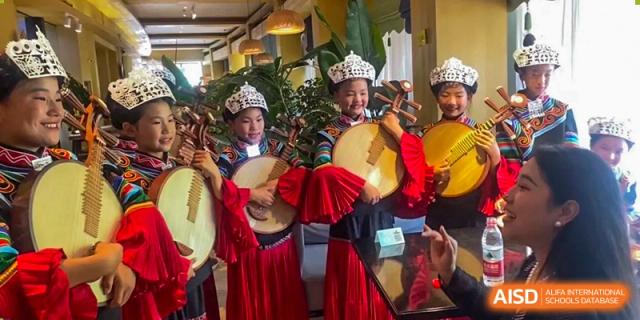 Two young students in traditional red and blue cultural attire play circular stringed instruments at Yew Wah International Education School of Guangzhou