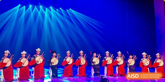 A group of performers in traditional red dresses play instruments on a stage under bright blue spotlights at Yew Wah International Education School of Guangzhou