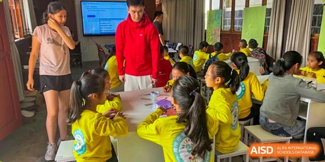 Students in yellow shirts gather around a table to work on a collaborative science or art project, guided by a teacher in a red hoodie at Beanstalk International Bilingual School Shunyi Campus.