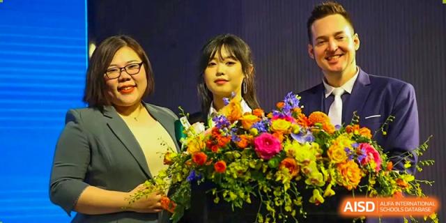 A student in a graduation gown and cap smiles while holding a large, vibrant bouquet of flowers alongside two faculty members at Beanstalk International Bilingual School Shunyi Campus.