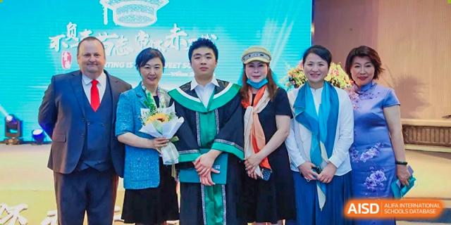 A graduate in a green and black gown stands proudly with family members and faculty, holding a bouquet of sunflowers after the ceremony at Beanstalk International Bilingual School Shunyi Campus.