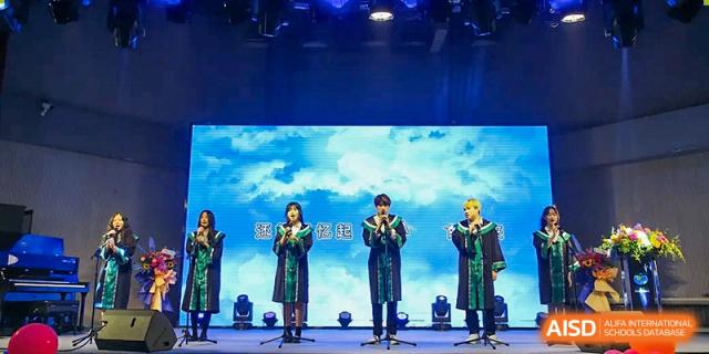 Four students in matching green and black graduation gowns perform a musical or vocal piece on stage during a school celebration at Beanstalk International Bilingual School Shunyi Campus.