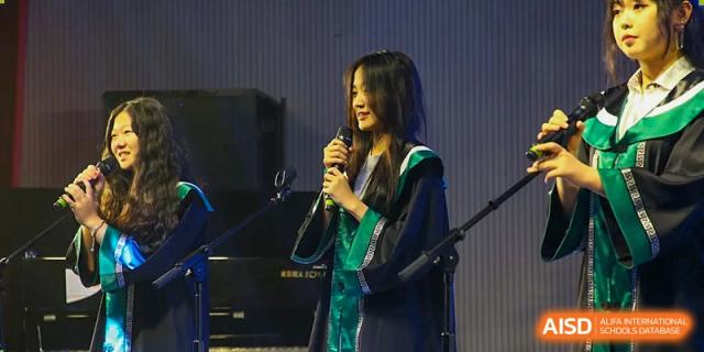 A student graduate speaks into a microphone on stage, delivering a speech or performing during a formal indoor academic event at Beanstalk International Bilingual School Shunyi Campus.