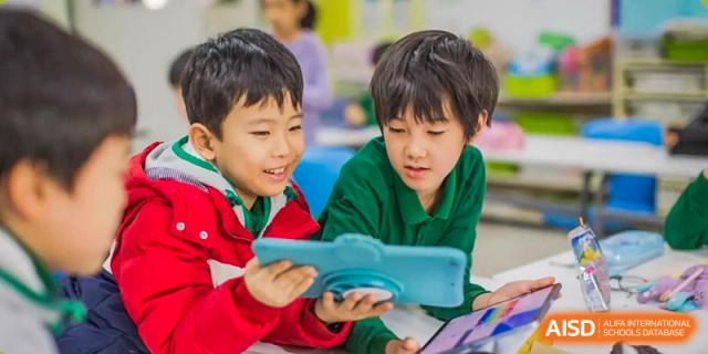 Two young students in red and green shirts smile while working together on a digital tablet, exploring educational apps in class at Beanstalk International Bilingual School Shunyi Campus.