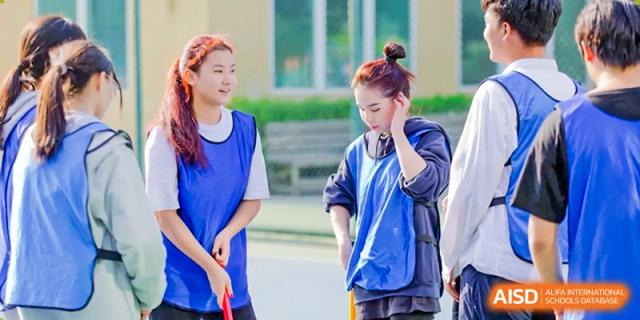 Two female students in blue sports vests stand on an outdoor court, holding floor hockey sticks and preparing for an active physical education match at Beanstalk International Bilingual School Shunyi Campus.