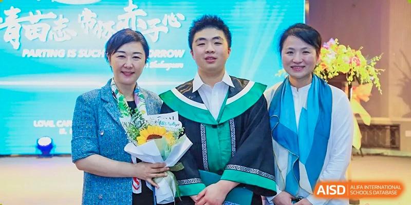 A proud graduating student in a green and black academic gown poses with two supportive family members while holding a beautiful bouquet of flowers at Beanstalk International Bilingual School Shunyi Campus.