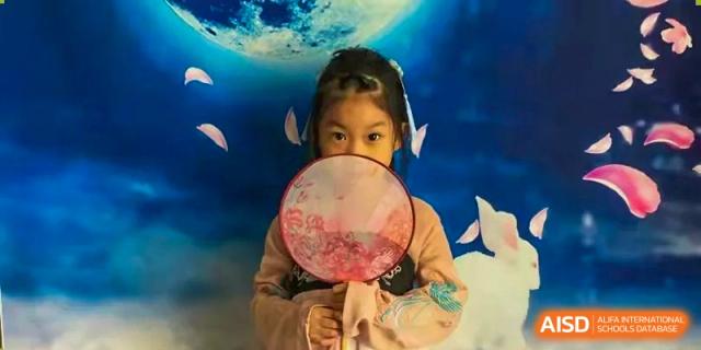 A young girl in a pink traditional dress holds a decorative circular fan in front of her face during a cultural celebration photoshoot at Beanstalk International Bilingual School Shunyi Campus.
