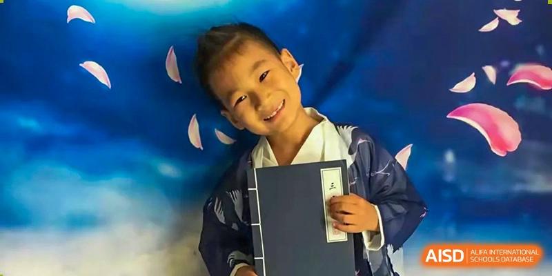 A young student smiles warmly while wearing traditional blue and white attire and holding a classic book against a celestial-themed backdrop at Beanstalk International Bilingual School Shunyi Campus.