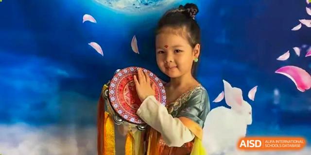 A young girl in ornate traditional clothing holds a red and gold tambourine, smiling during a creative arts performance or cultural event at Beanstalk International Bilingual School Shunyi Campus.
