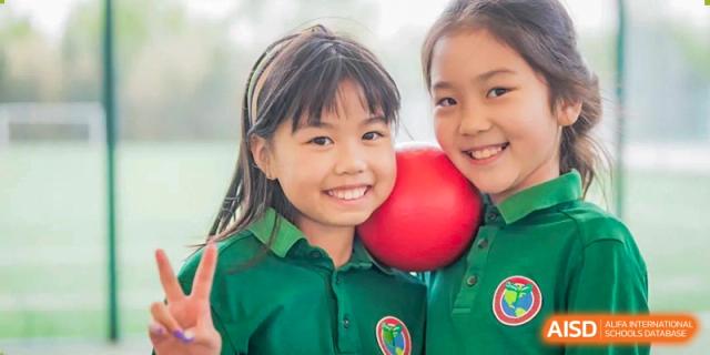 Two young friends in green polo shirts smile brightly while holding a red ball and making peace signs during a fun break time on the playground at Beanstalk International Bilingual School Shunyi Campus.