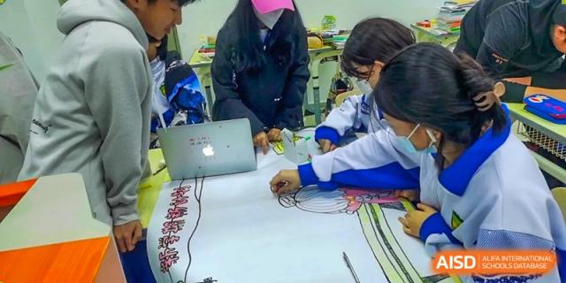 Focused students collaborate on a large artistic drawing spread across a table while utilizing a laptop for creative reference in a bright classroom at Beijing LuHe International Academy.