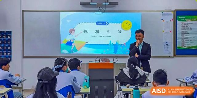 A male teacher in a formal suit stands by a digital screen and leads an engaging presentation for students seated at their desks in a modern classroom at Beijing LuHe International Academy.
