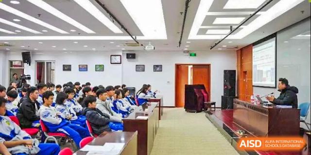 A large group of students in blue and white school uniforms sits in a tiered lecture hall and listens attentively to an academic presentation at Beijing LuHe International Academy.