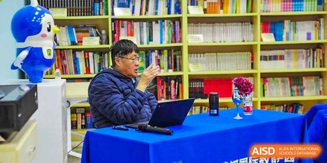 An educator sits at a blue table with a laptop and speaks to students in a library setting filled with bookshelves to facilitate a deep discussion at Beijing LuHe International Academy.