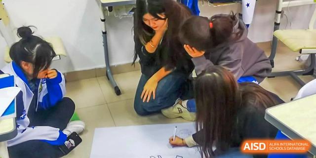 Several students sit on the floor together to brainstorm and sketch out creative ideas on a large white poster during a collaborative group project at Beijing LuHe International Academy.