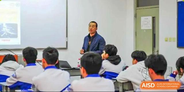 A teacher stands at the front of the room and gestures while delivering an informative lecture to a class of students wearing matching blue uniforms at Beijing LuHe International Academy.