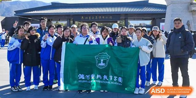 A group of students in blue school tracksuits stands together outdoors while proudly holding a large green flag featuring the school crest at Beijing LuHe International Academy.