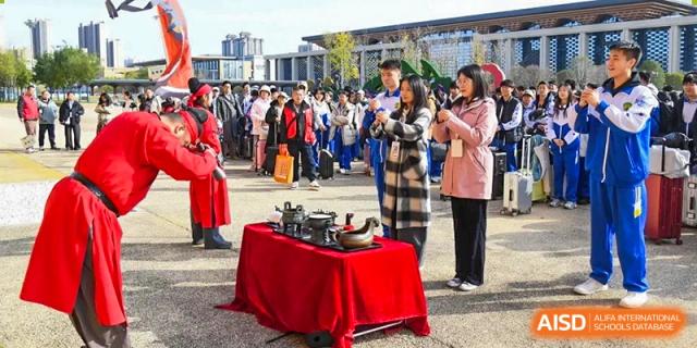 Students participate in a traditional tea ceremony demonstration outdoors with a red table and ritual teapots during a cultural exchange event at Beijing LuHe International Academy.