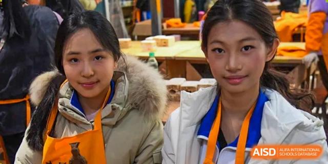 Two female students in warm winter coats and orange lanyards smile for a close-up portrait while participating in a hands-on workshop at Beijing LuHe International Academy.