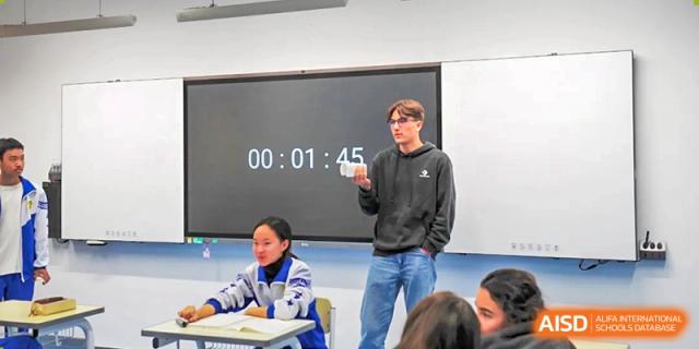 A student in a black hoodie stands before a digital timer on a screen and leads a presentation for a small group of classmates at Beijing LuHe International Academy.