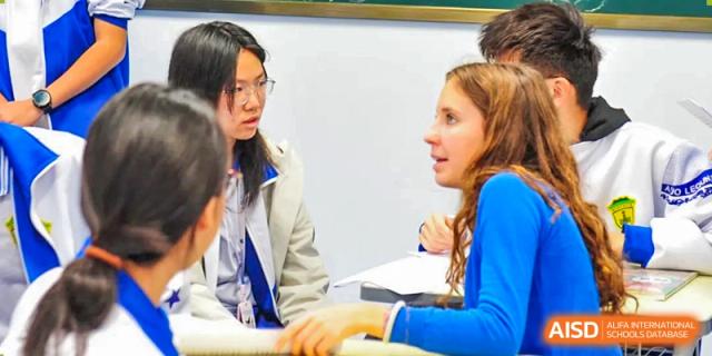 Students in blue school jackets and casual wear participate in a peer-to-peer learning session by discussing notes and sharing feedback at Beijing LuHe International Academy.