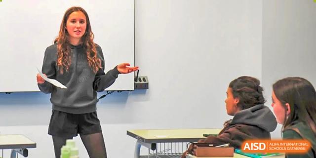 A female student stands confidently at the front of a classroom and uses hand gestures while delivering a presentation to her seated classmates at Beijing LuHe International Academy.