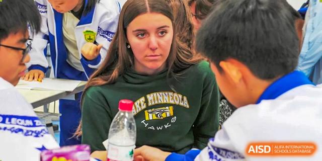 A focused female student wearing a green sweatshirt listens intently to a peer during a collaborative classroom activity designed to foster teamwork at Beijing LuHe International Academy.
